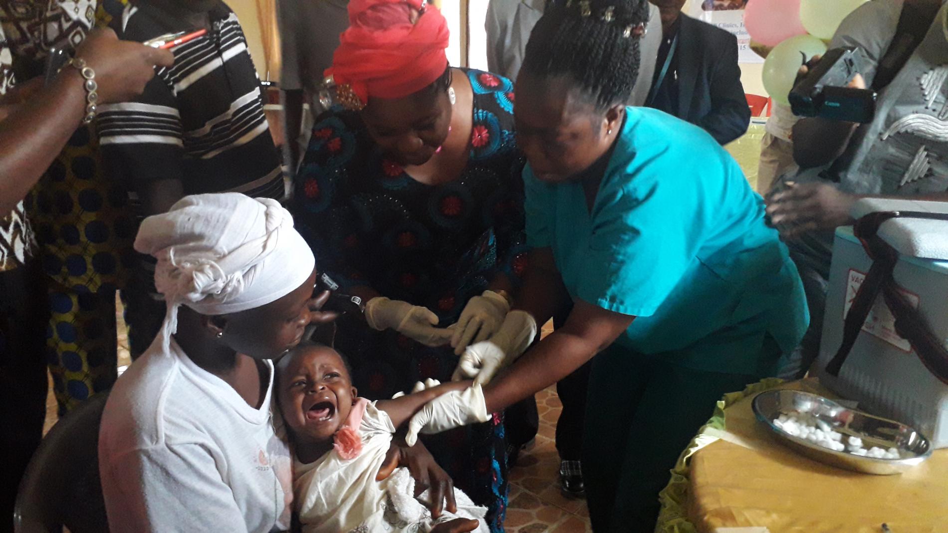 Vaccination of the first child by a healthcare worker, assisted by Amb. Julie Endee during the MCV2 launch in Bomi County, Liberia