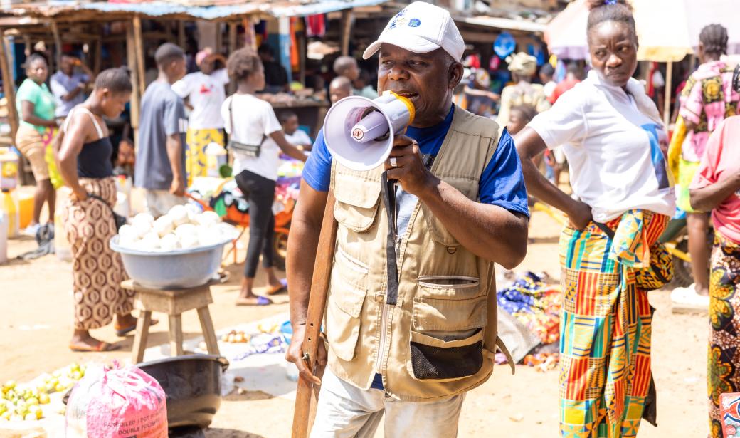 Community mobiliser sharing information in the marketplace on the polio vaccination campaign, with emphasis on the benefits of the vaccine, surrounded by curious listeners.